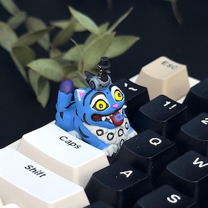 May include: A blue and white cartoon cat keycap with yellow eyes and a small hat sits on a keyboard. The keyboard has black and white keys. The keycap is on the "Caps" key. The background is dark with some green leaves.