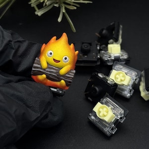 May include: A vibrant artisan keycap depicting a smiling fire character holding a log. The keycap is yellow, orange, and brown. Several mechanical keyboard switches are also visible.