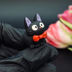 May include: A small, black cat figurine with a red bow tie and large white eyes. The cat has purple accents on its ears. The figurine is held in a black gloved hand, with pink and white tulips in the background.