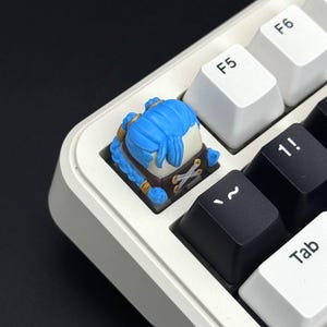 Op de afbeelding: Een aangepaste artisanale mechanische toetsenbordtoets met een gebeeldhouwd figuur met felblauw haar, gevlochten staartjes en een bruine outfit met witte veterdetails, geïnstalleerd op een wit en zwart mechanisch toetsenbord bij de F5-toets.