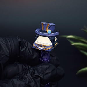May include: A custom artisan keycap with a face design and a top hat. The keycap is mainly purple, with gold and blue details. The face has a white mouth and chin, and black hair. The keycap is held by a gloved hand.