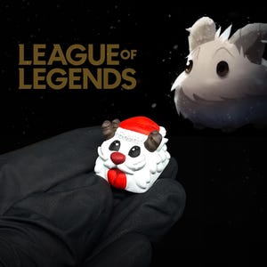 May include: A white and red Santa hat Poro keycap from League of Legends, held in a gloved hand. The keycap has a red nose and mouth, with small brown antlers. The League of Legends logo is in gold on a black background.