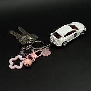 Peut inclure: Un porte-clés avec une voiture jouet blanche, un porte-clés argenté, des clés et des breloques roses. Les breloques comprennent une étoile, une cloche et un fermoir. La voiture a des roues noires et un intérieur rouge.