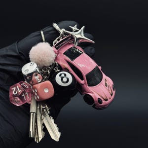 May include: A pink car keyring with a star charm, dice, a fluffy pink ball, and an 8-ball charm. The keyring also holds several keys. The car has the word "PORSCHE" printed on it.