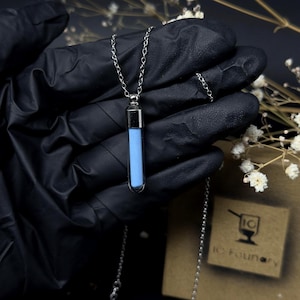 Op de afbeelding: Zilverkleurige ketting met een kleine, rechthoekige vial-hanger gevuld met een lichtblauwe substantie. De vial hangt aan een fijne ketting. De achtergrond is donker, met bloemmotieven.