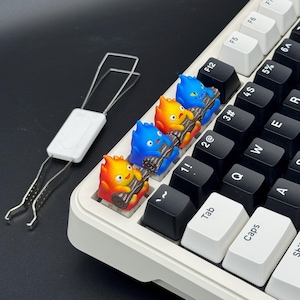 Op de afbeelding: Een mechanisch toetsenbord met aangepaste keycaps met kleurrijke vuurachtige karakters die boomstammen vasthouden. De keycaps zijn rood, oranje en blauw. Er is ook een keycap-trekker aanwezig.