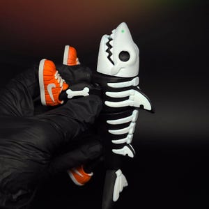 May include: A black and white shark skeleton figurine with a white jaw and teeth. The figurine is holding miniature orange and white trainers. The trainers have a white swoosh logo on the side.