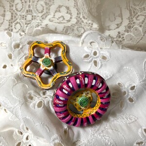 May include: Two decorative, hand-painted knobs. One is a star shape with yellow, red, and green accents. The other is a circular shape with pink, yellow, and purple details. Both have a green spiral in the center.