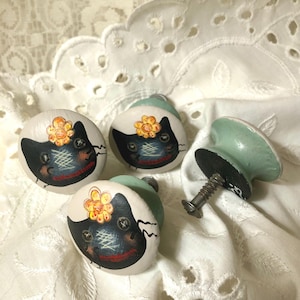 May include: Four decorative cabinet knobs with a whimsical cat design. The knobs are painted with a black cat face, a yellow flower, and a red smile. One knob is detached, showing the screw and the green base.