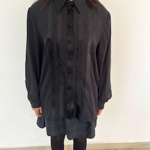 Può includere: Camicia nera abbottonata con dettagli a righe verticali sul davanti. La camicia ha un colletto classico e maniche lunghe. La modella indossa una gonna a quadri e collant neri.