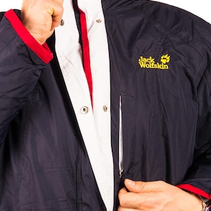 May include: Navy blue jacket with white and red accents. The jacket features a white collar and a red trim on the cuffs. The Jack Wolfskin logo is visible on the right chest. The jacket has a zipper closure and snap buttons.