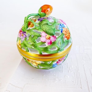 May include: A round porcelain trinket box featuring an intricate openwork lattice design of green leaves and colorful painted flowers. The lid is topped with a small orange fruit-shaped handle and the center is accented with a gold band.