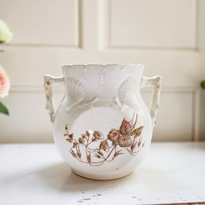May include: A white ceramic vase with two handles and a scalloped rim. The vase features a floral design in brown and pink tones. The handles have gold accents. The vase is sitting on a white surface.