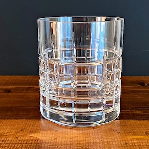 May include: A clear, lowball glass with a grid-like pattern etched into the exterior. The glass sits on a wooden surface, reflecting light. The design gives the glass a classic, textured appearance, suitable for serving spirits or cocktails.
