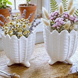 May include: Two white ceramic planters with a scalloped rim and three small feet. One planter holds yellow and purple flowers, the other holds pink flowers and fluffy beige stems. The planters sit on a wooden surface.