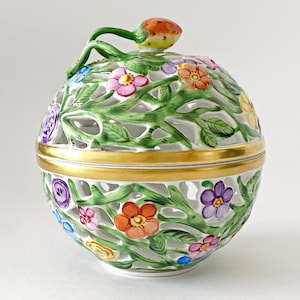 May include: A white, ceramic decorative lidded box with a floral design. The box features a gold band around the middle and is adorned with colorful flowers and green leaves. The lid is topped with a bud-shaped finial.