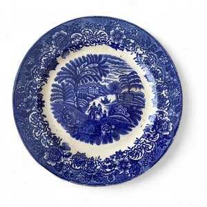 May include: A round ceramic plate featuring a blue and white transferware pattern. The center depicts a landscape with trees, figures, and a building, while the wide border is decorated with a dense floral motif of various blossoms and leaves.