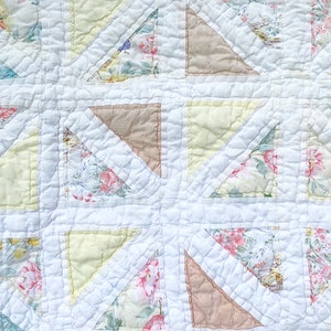 May include: A quilted textile featuring a geometric pattern of triangles separated by white fabric strips. The design incorporates various floral prints in shades of pink, blue, and yellow, alongside solid light brown and pale yellow triangular patches.
