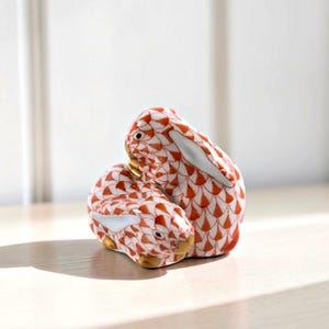 May include: A pair of ceramic rabbit figurines. The rabbits are white with a red and orange fishnet pattern. They have white ears and gold accents on their faces. The figurines are displayed on a light-colored surface.