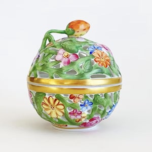 May include: A decorative, lidded ceramic jar with a floral design. The jar is spherical, with a pierced green exterior adorned with colorful flowers. A gold band encircles the middle, and a small orange fruit tops the lid. The jar is likely used for storing small items.
