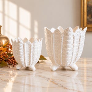 May include: Two white ceramic planters with a textured, leaf-like exterior and scalloped rims. Each planter features three small, rounded feet at the base. The vessels have a matte finish and a vertical ribbed pattern that mimics natural foliage.