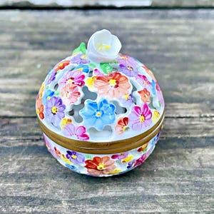 May include: A decorative, floral-patterned trinket box. The box is round with a lid and is adorned with colorful, hand-painted flowers in shades of pink, purple, blue, and orange. A white flower sits atop the lid. The box has a gold-colored band around the middle.