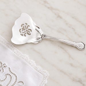 May include: A silver-toned metal cake server featuring a wide, fan-shaped blade with a central cut-out scrollwork design. The handle is slender with a curved, scalloped edge and a small engraved monogram detail at the very end of the handle.