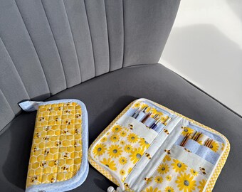 May include: Two fabric needle cases, one closed and one open, rest on a gray chair. The closed case features a yellow honeycomb pattern with bees, while the open case displays a sunflower design. Both cases have blue trim and pearl zipper pulls.