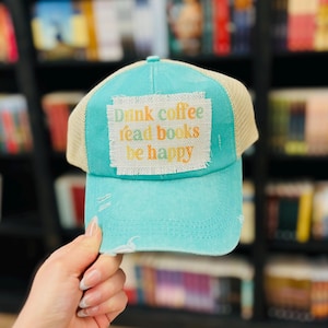 Puede incluir: Una gorra de camionero azul turquesa con una malla beige en la parte posterior. La gorra tiene un aspecto desgastado y presenta un parche con el texto "Drink coffee read books be happy" en letras de colores.