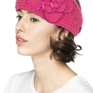 K&ouml;nnte beinhalten: Ein magentafarbenes Strickstirnband mit einer gro&szlig;en, mit Pailletten besetzten Blumenapplikation. Die Blume ist mittig auf dem Stirnband angebracht und hat mehrere Bl&uuml;tenblattschichten. Das Stirnband besteht aus dickem, strukturiertem Garn.