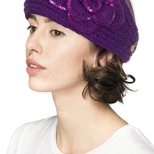 K&ouml;nnte beinhalten: Ein lila Strickbarett mit einer gro&szlig;en, dekorativen Blume auf der Vorderseite. Die Blume ist mit Pailletten verziert und hat ein mehrschichtiges Design. Auf der Seite des Baretts befindet sich ein Knopf. Ein modisches Accessoire.