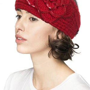 K&ouml;nnte beinhalten: Ein rotes Stirnband aus Strick mit einer gro&szlig;en, mit Pailletten besetzten Blume. Das Stirnband ist aus einem dicken, strukturierten Garn gefertigt und wird auf dem Kopf getragen. Die Blume ist das zentrale Dekorationselement.