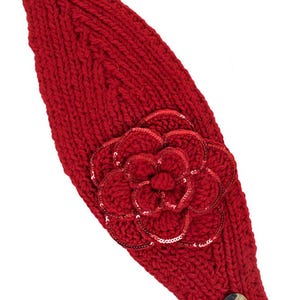 K&ouml;nnte beinhalten: Ein leuchtend rotes Stirnband aus Strick mit einer gro&szlig;en, paillettenbesetzten Blume. Das Stirnband hat ein strukturiertes Strickdesign und ist mit zwei dekorativen Kn&ouml;pfen verziert. Das Wort "Red" ist unten rechts aufgedruckt.