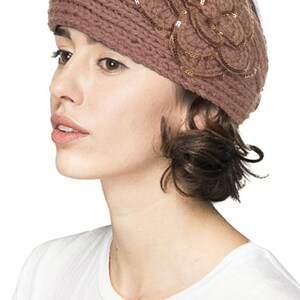 K&ouml;nnte beinhalten: Gestricktes Stirnband in ged&auml;mpftem Mauve, verziert mit einem Blumendesign mit goldenen Pailletten. Das Stirnband besteht aus dickem, strukturiertem Garn und wird auf dem Kopf getragen.