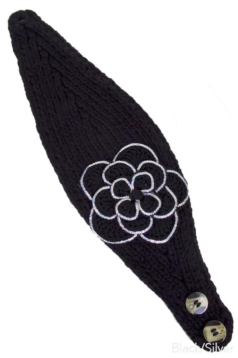 K&ouml;nnte beinhalten: Schwarzes Stirnband aus Strick mit einer gro&szlig;en, silberfarben umrandeten Blumenapplikation. Das Stirnband hat zwei dekorative Kn&ouml;pfe und den Text "Black/Silver". Ein stilvolles Accessoire.