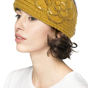 K&ouml;nnte beinhalten: Senfgelbes Strickstirnband mit einer gro&szlig;en, dekorativen Blume. Die Blume ist mit goldfarbenen Pailletten verziert. Das Stirnband ist ein stilvolles Accessoire, um die Ohren warm zu halten. Das Model tr&auml;gt ein wei&szlig;es T-Shirt.