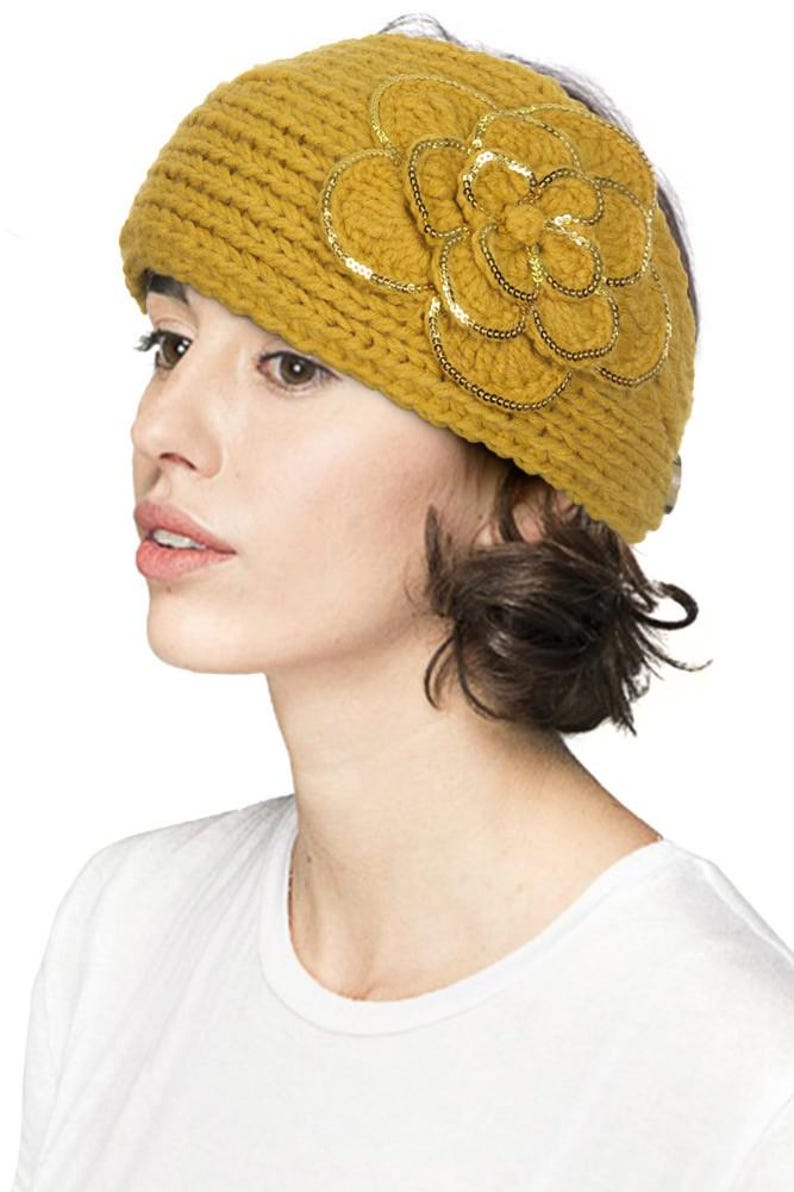 K&ouml;nnte beinhalten: Senfgelbes Strickstirnband mit einer gro&szlig;en, dekorativen Blume. Die Blume ist mit goldfarbenen Pailletten verziert. Das Stirnband ist ein stilvolles Accessoire, um die Ohren warm zu halten. Das Model tr&auml;gt ein wei&szlig;es T-Shirt.