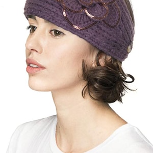 K&ouml;nnte beinhalten: Gestricktes lila Stirnband mit gro&szlig;em Blumenapplikation. Die Blume ist mit Pailletten verziert. Das Stirnband wird von einer Person getragen, die ein wei&szlig;es T-Shirt tr&auml;gt.