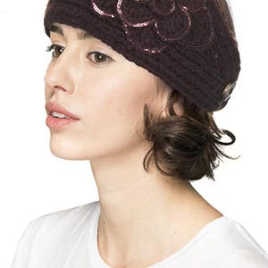 K&ouml;nnte beinhalten: Dunkelbraunes Strickstirnband mit gro&szlig;em Blumenapplikation. Die Blume ist mit Pailletten verziert und hat ein Knopfdetail an der Seite. Das Stirnband wird von einer Person getragen, die ein wei&szlig;es T-Shirt tr&auml;gt.