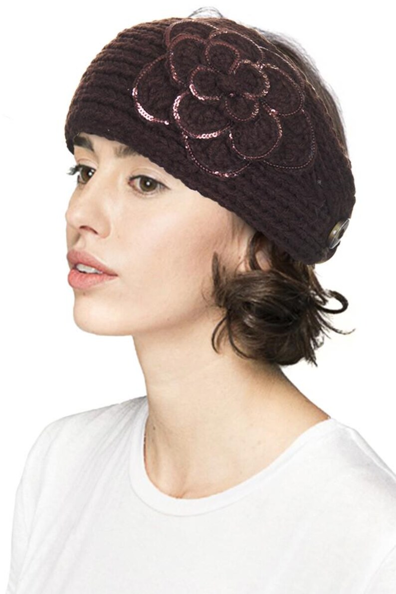 K&ouml;nnte beinhalten: Dunkelbraunes Strickstirnband mit gro&szlig;em Blumenapplikation. Die Blume ist mit Pailletten verziert und hat ein Knopfdetail an der Seite. Das Stirnband wird von einer Person getragen, die ein wei&szlig;es T-Shirt tr&auml;gt.