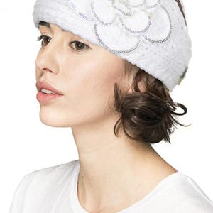 K&ouml;nnte beinhalten: Ein wei&szlig;er Strickhut mit einer gro&szlig;en, dekorativen Blume an der Seite. Die Blume ist mit silbernen Pailletten verziert. Die Person tr&auml;gt ein wei&szlig;es T-Shirt.