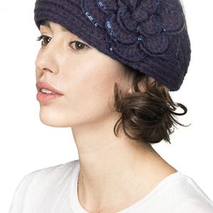 K&ouml;nnte beinhalten: Ein marineblauer Strickbarett mit einer gro&szlig;en, dekorativen Blume. Die Blume ist mit Pailletten verziert und hat die gleiche Farbe wie der Hut. Das Barett wird von einer Person getragen, die ein wei&szlig;es T-Shirt tr&auml;gt.