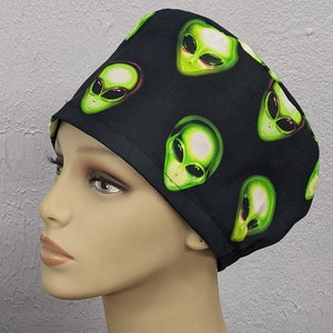 May include: Black surgical cap featuring a repeating pattern of green alien faces. The alien faces have large black eyes and are outlined in red. The cap is made of a soft, dark fabric and is designed to cover the head.