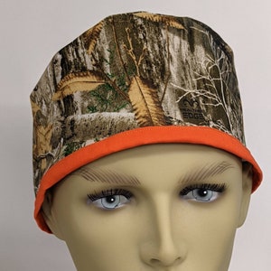 May include: A camouflage patterned scrub hat with an orange trim. The hat has the text "Mountain Edge" embroidered on the side.