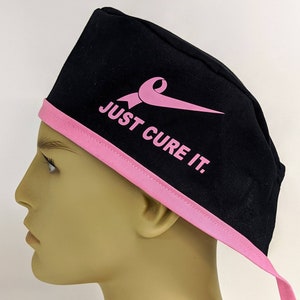 May include: Black scrub hat with a pink ribbon and the text "JUST CURE IT."