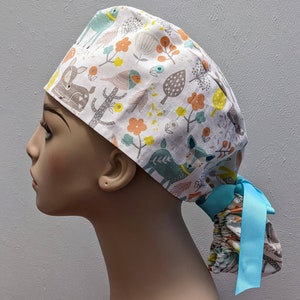 May include: A white scrub hat with a colorful pattern of deer, birds, trees, and flowers. The hat has a blue ribbon tie.