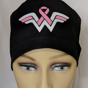 May include: Black surgical scrub cap featuring a pink ribbon and a white and pink Wonder Woman logo. The cap is made of a dark fabric and is designed to be worn in a medical setting. The Wonder Woman logo is prominently displayed on the front.