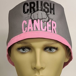 May include: Gray scrub cap with pink trim and ties. The cap features the words "CRUSH CANCER" in black and pink, with a graphic of a fist breaking through a surface.