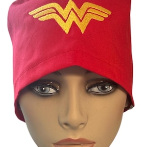 May include: Red scrub cap with a gold Wonder Woman logo embroidered on the front. The cap is made of a soft fabric and is designed to fit comfortably. This cap is a great accessory for healthcare professionals.