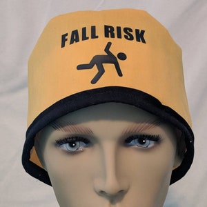 May include: Yellow cap with black trim and the words "FALL RISK" in black. A black silhouette of a person falling is also on the cap. The cap is worn by a mannequin.